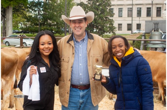 Local television news reporters Kayla Thompson with WJTV and Morgan Harris with WLBT competed in the first milking contest. Kayla Thompson won the round after milking a total of 1.5 cups of milk from Sophia the cow. Pictured L-R: Morgan Harris, WLBT Reporter; Ag Commissioner Andy Gipson; and Kayla Thompson, WJTV Reporter.